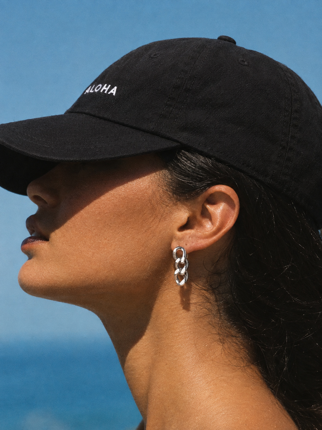 A close-up side profile of a woman wearing a black baseball cap with 'ALOHA' embroidered in white on the front, set against a blue sky and ocean background. She is also wearing a silver link earring.