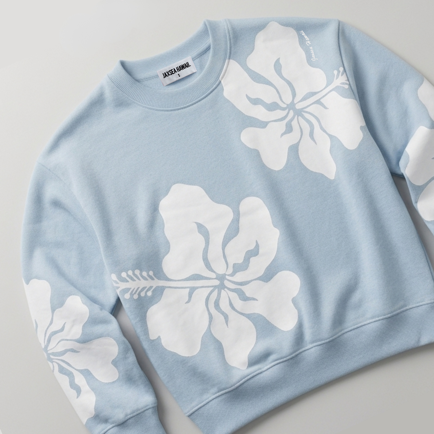 A light blue crewneck sweatshirt with large white hibiscus floral prints on the front and sleeves, shown on a neutral light gray background.