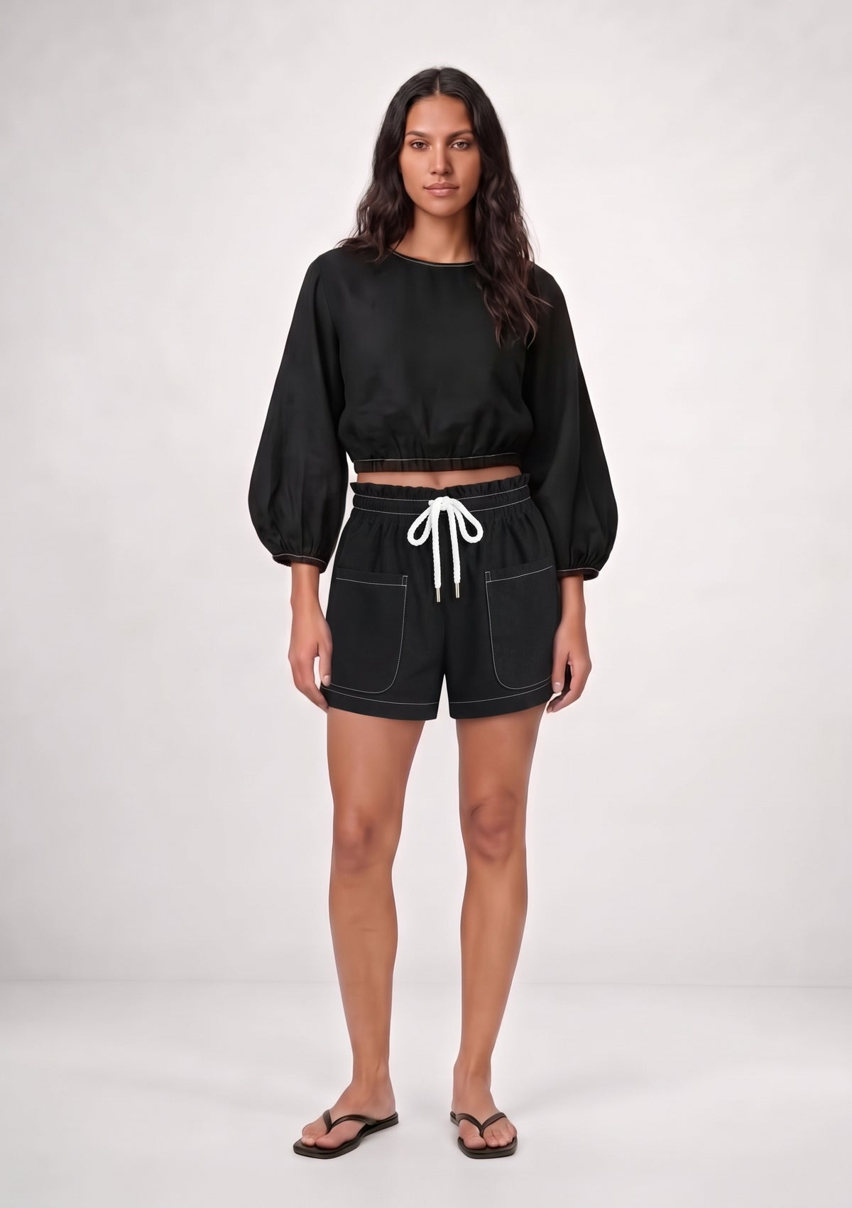 A woman modeling a black two-piece outfit consisting of a long-sleeve cropped top with balloon sleeves and matching black shorts with a white drawstring, standing against a light gray studio background.