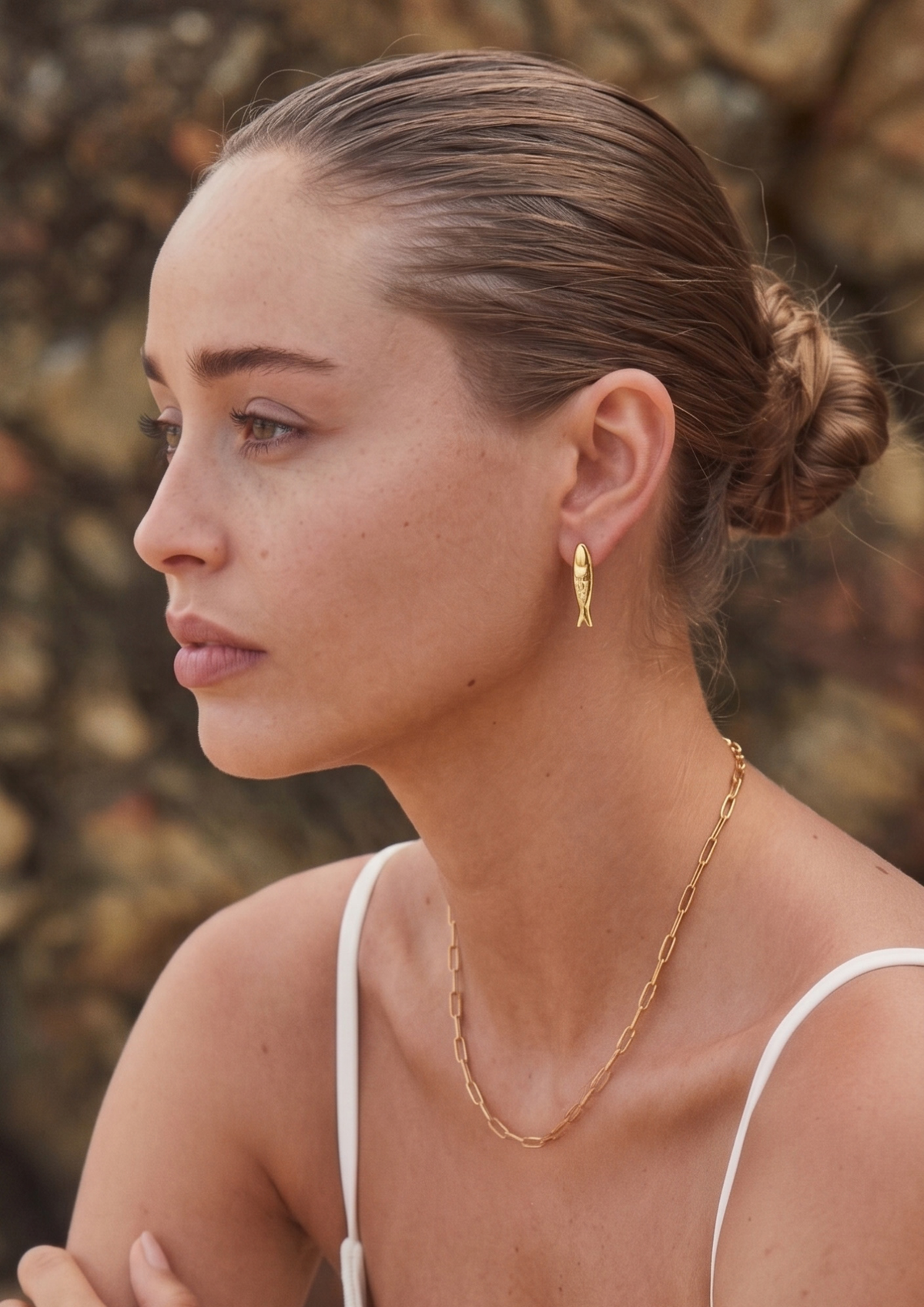 A woman in profile wearing a delicate gold chain necklace and a small gold hoop earring, photographed outdoors against a blurred rocky background.