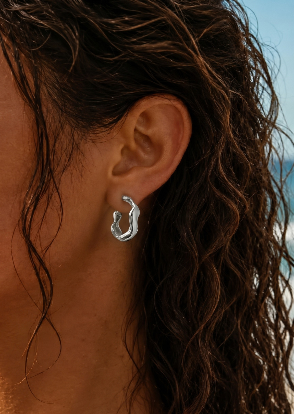 A close-up of a person's ear wearing a small, polished silver huggie hoop earring, with wet brown hair and a blurred outdoor background.